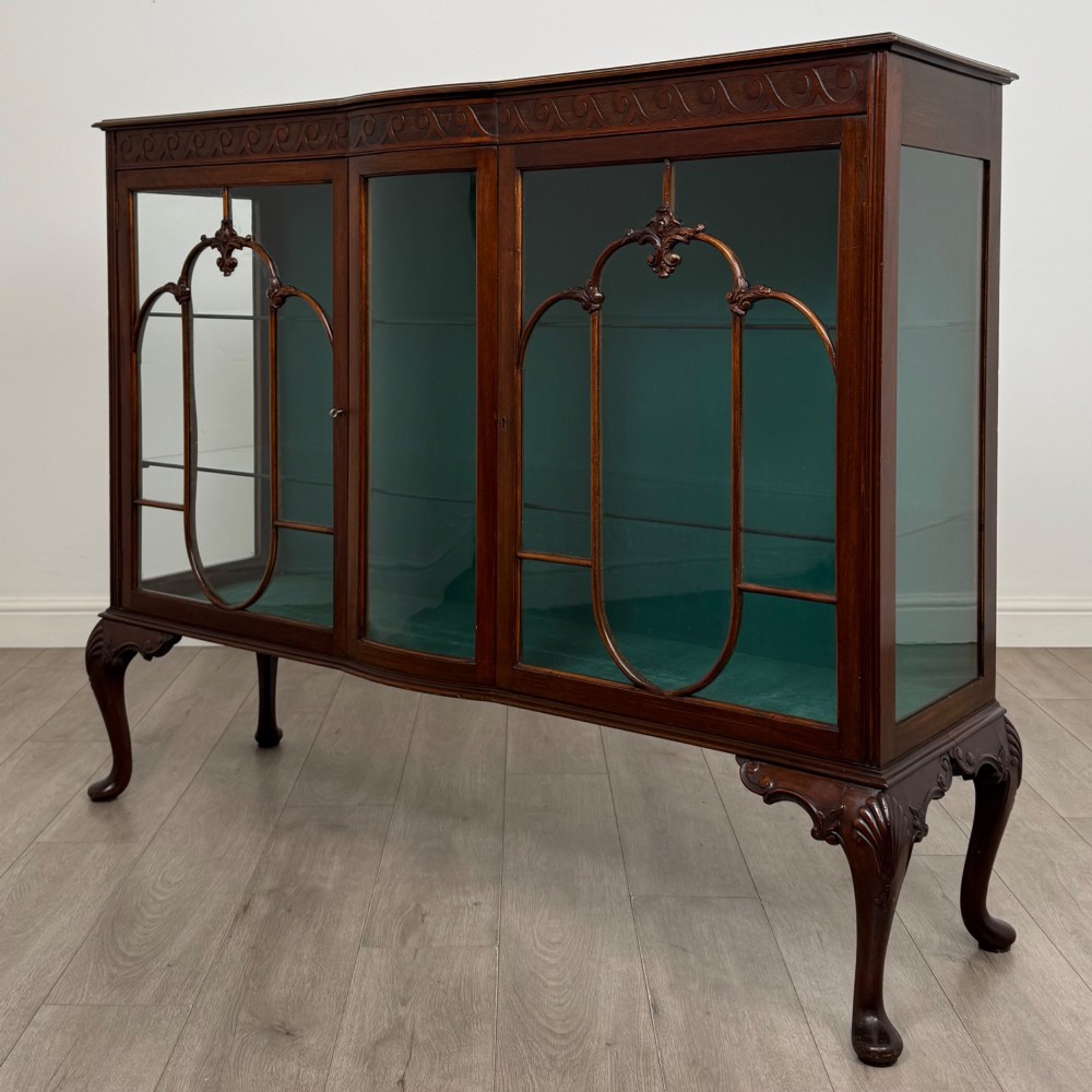 An early twentieth-century mahogany display cabinet featuring three glazed doors with curved astragal glazing bars, a carved top frieze, green fabric interior, and cabriole legs. View III