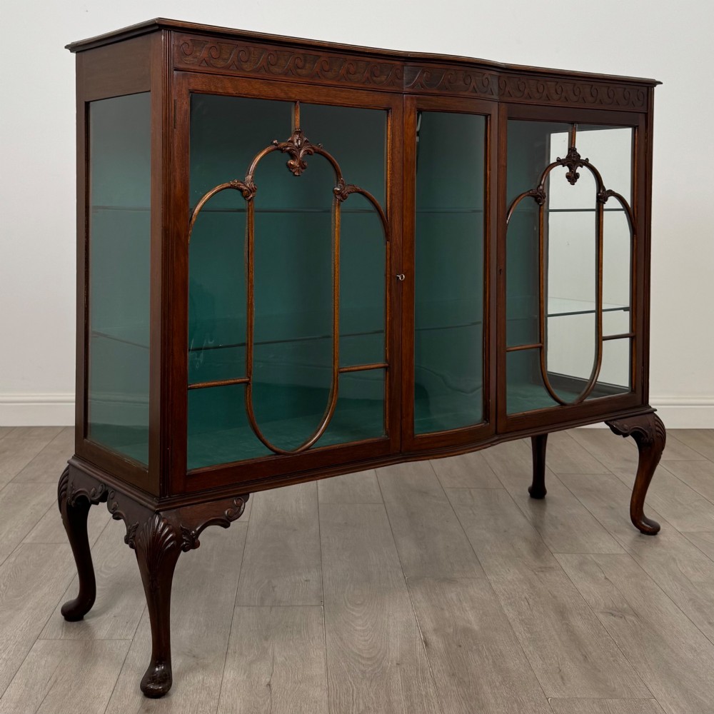 An early twentieth-century mahogany display cabinet featuring three glazed doors with curved astragal glazing bars, a carved top frieze, green fabric interior, and cabriole legs. View IX