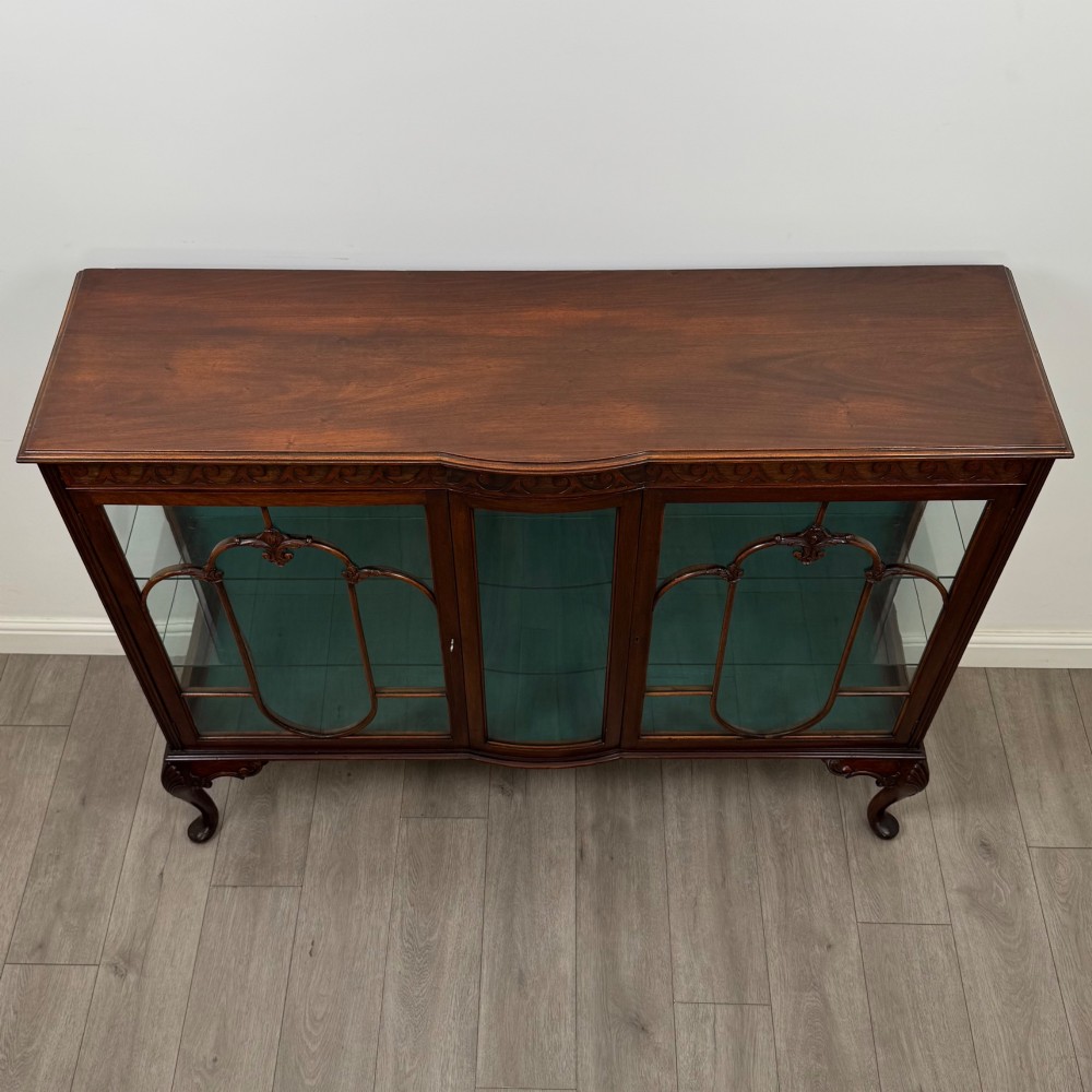 An early twentieth-century mahogany display cabinet featuring three glazed doors with curved astragal glazing bars, a carved top frieze, green fabric interior, and cabriole legs. View X