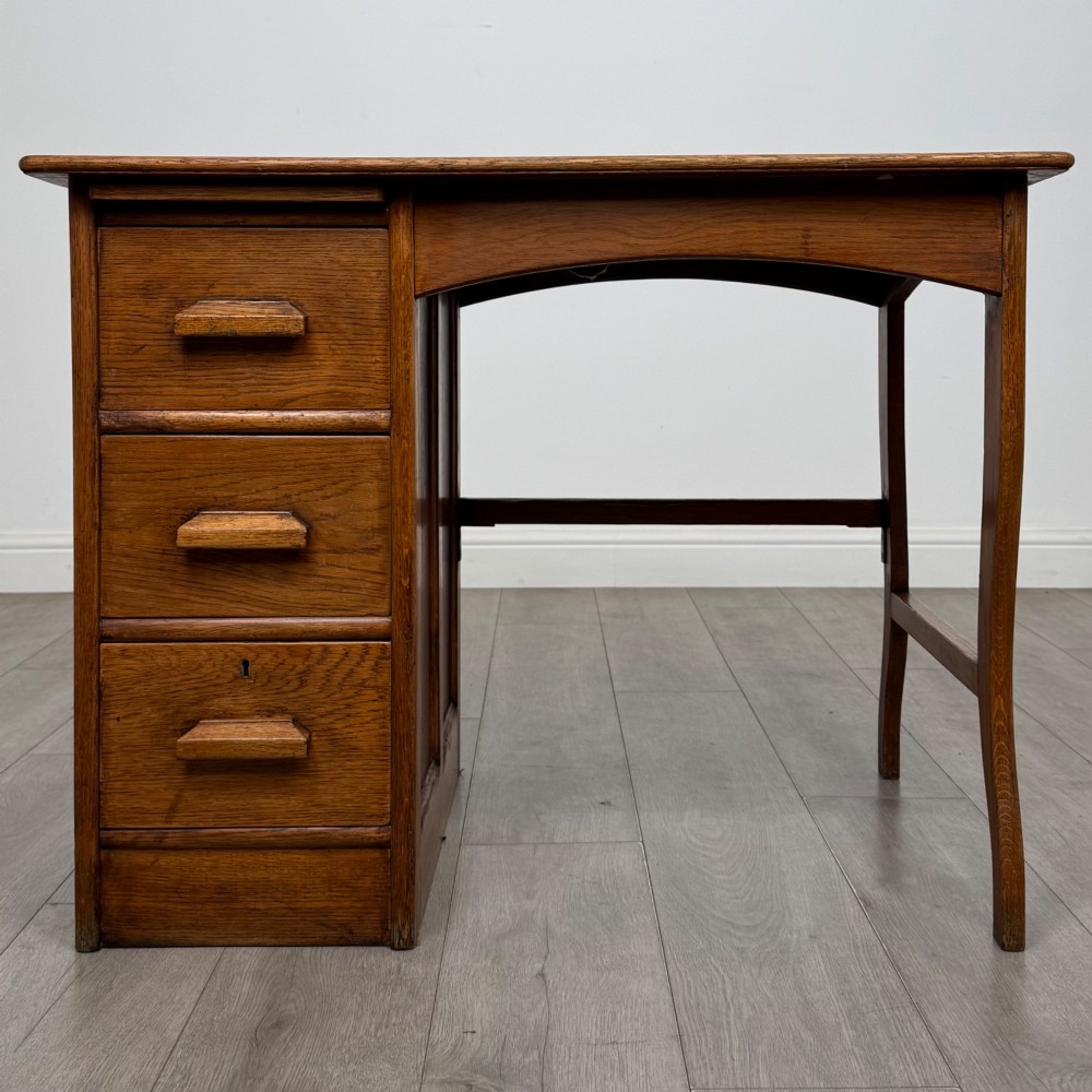 An Edwardian single pedestal oak desk with three stacked drawers on the left, integrated wood pulls, and an open kneehole section supported by slim right-side legs.
