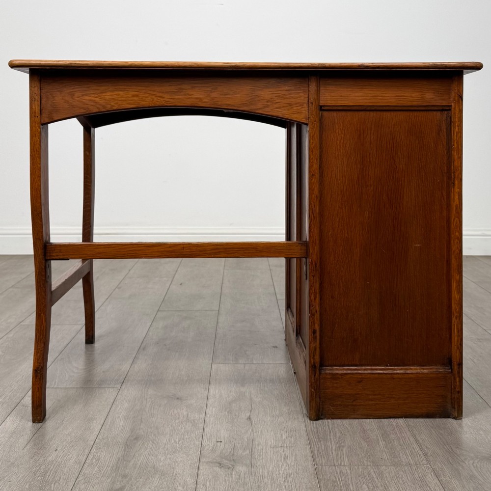 An Edwardian single pedestal oak desk with three stacked drawers on the left, integrated wood pulls, and an open kneehole section supported by slim right-side legs. View V