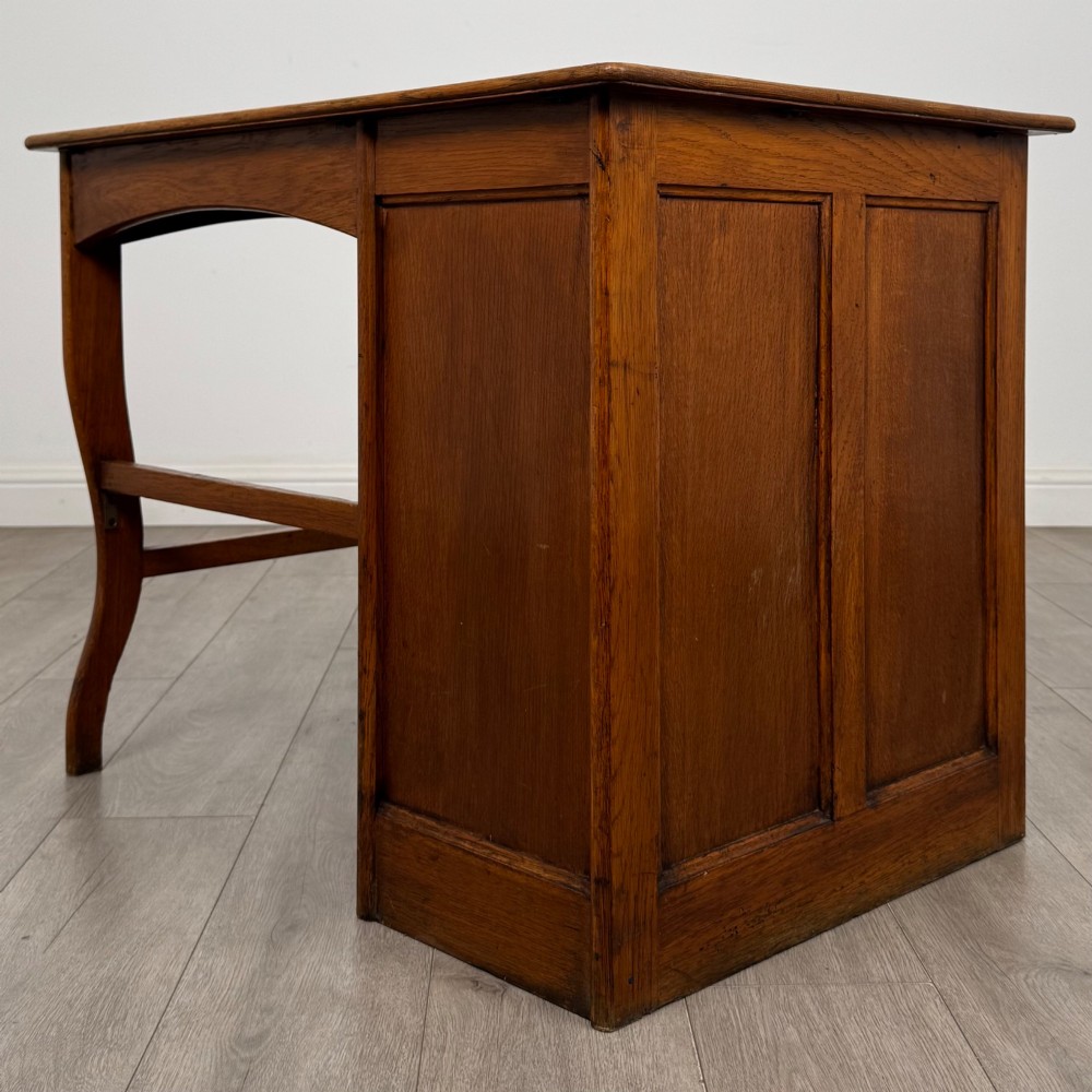 An Edwardian single pedestal oak desk with three stacked drawers on the left, integrated wood pulls, and an open kneehole section supported by slim right-side legs. View VI