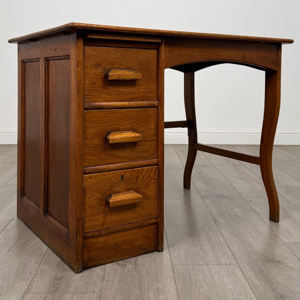 An Edwardian single pedestal oak desk with three stacked drawers on the left, integrated wood pulls, and an open kneehole section supported by slim right-side legs. View VIII