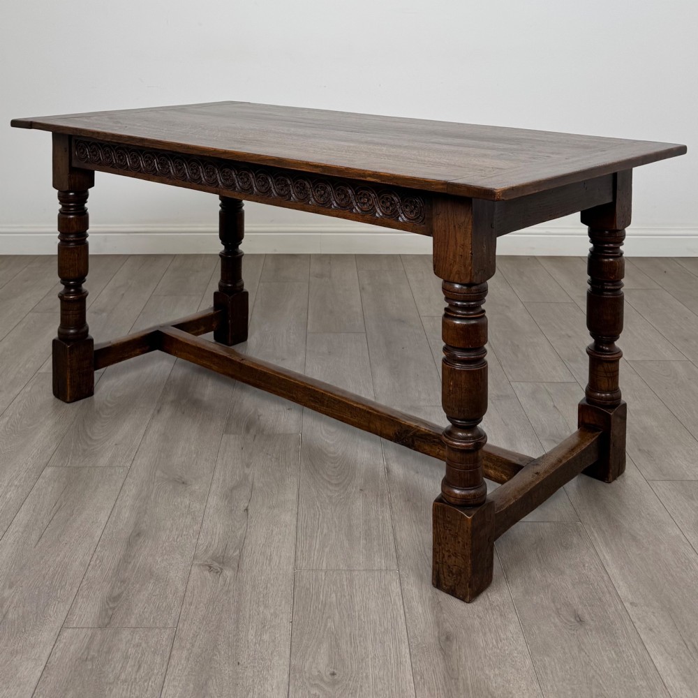 A dark carved oak refectory dining table featuring a carved frieze apron, turned baluster legs, and an H-stretcher base on light timber flooring.