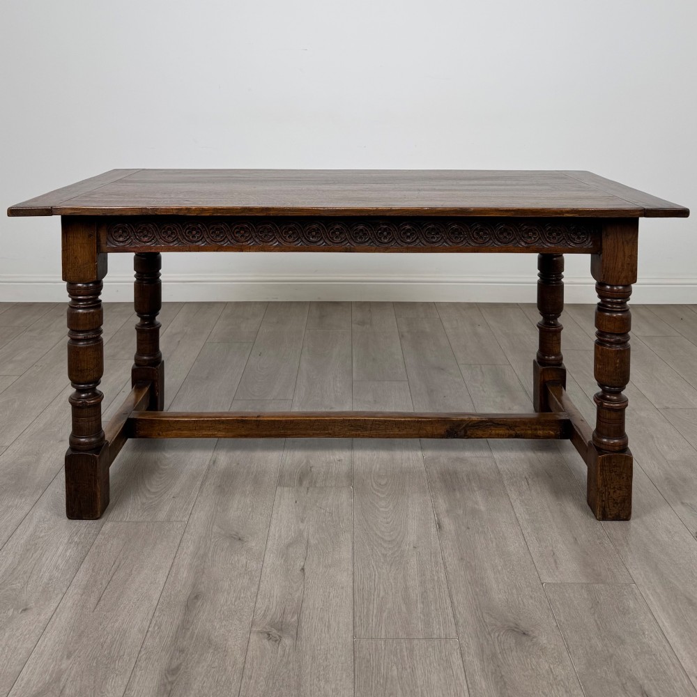 A dark carved oak refectory dining table featuring a carved frieze apron, turned baluster legs, and an H-stretcher base on light timber flooring. View II
