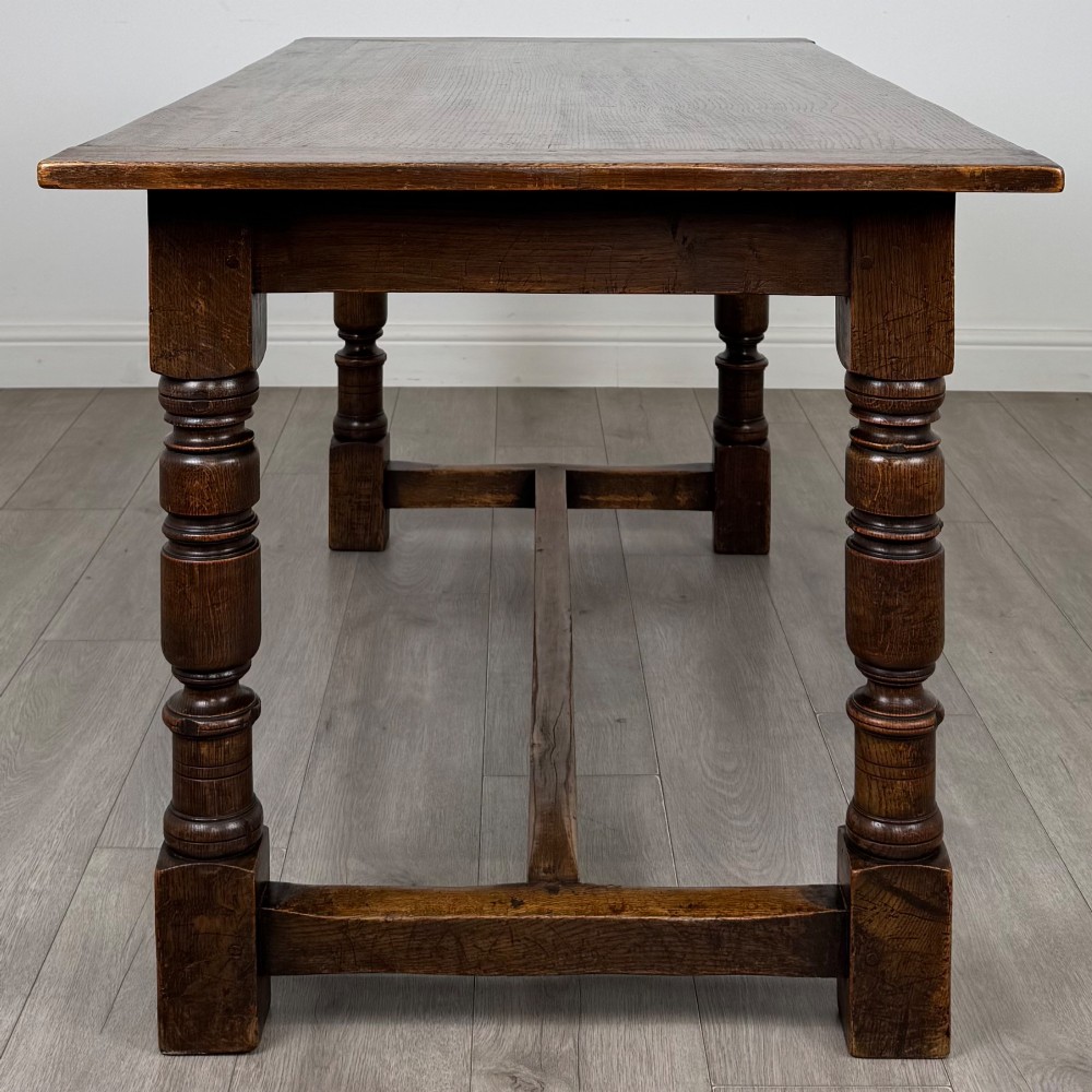 A dark carved oak refectory dining table featuring a carved frieze apron, turned baluster legs, and an H-stretcher base on light timber flooring. View III