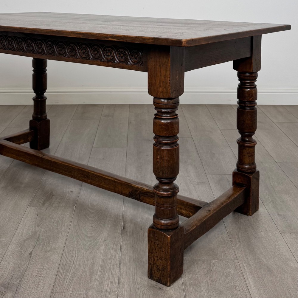 A dark carved oak refectory dining table featuring a carved frieze apron, turned baluster legs, and an H-stretcher base on light timber flooring. View V