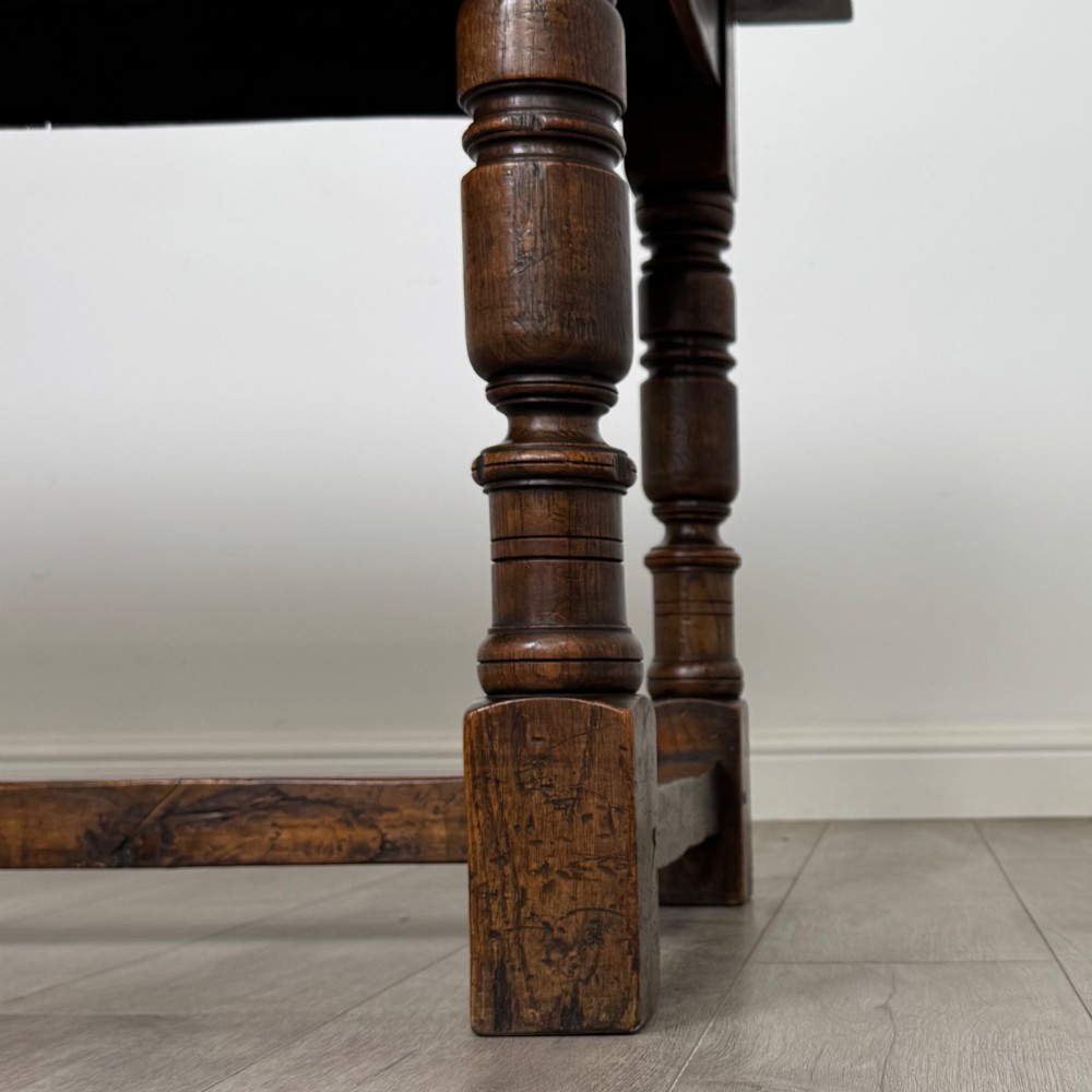 A dark carved oak refectory dining table featuring a carved frieze apron, turned baluster legs, and an H-stretcher base on light timber flooring. View VI
