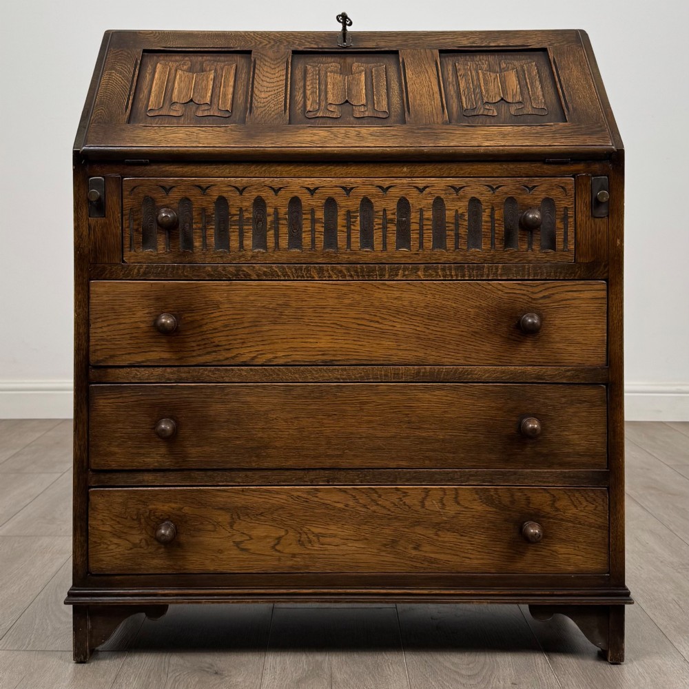A dark oak writing bureau with a slanted fall-front upper section featuring three carved linenfold panels above four drawers with turned wooden knob handles.