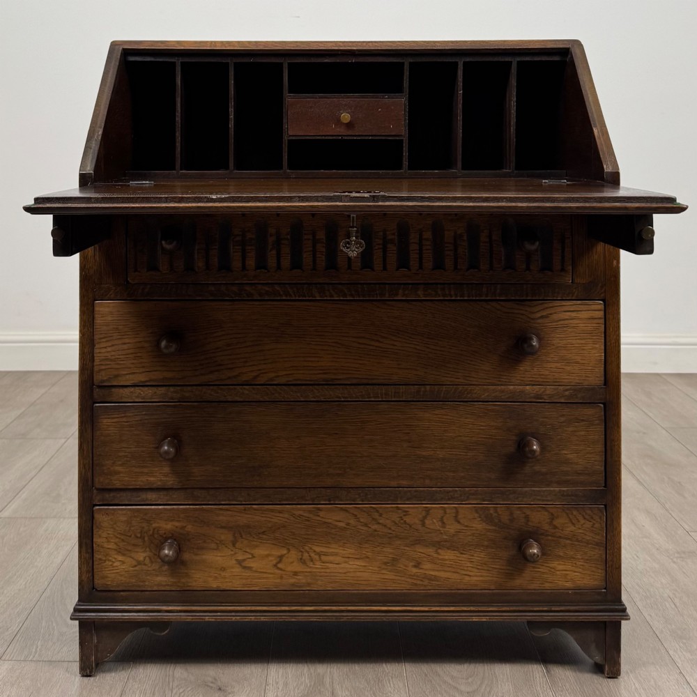 A dark oak writing bureau with a slanted fall-front upper section featuring three carved linenfold panels above four drawers with turned wooden knob handles. View II