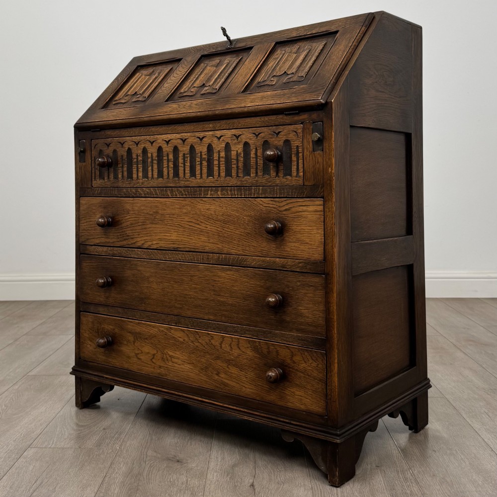 A dark oak writing bureau with a slanted fall-front upper section featuring three carved linenfold panels above four drawers with turned wooden knob handles. View III