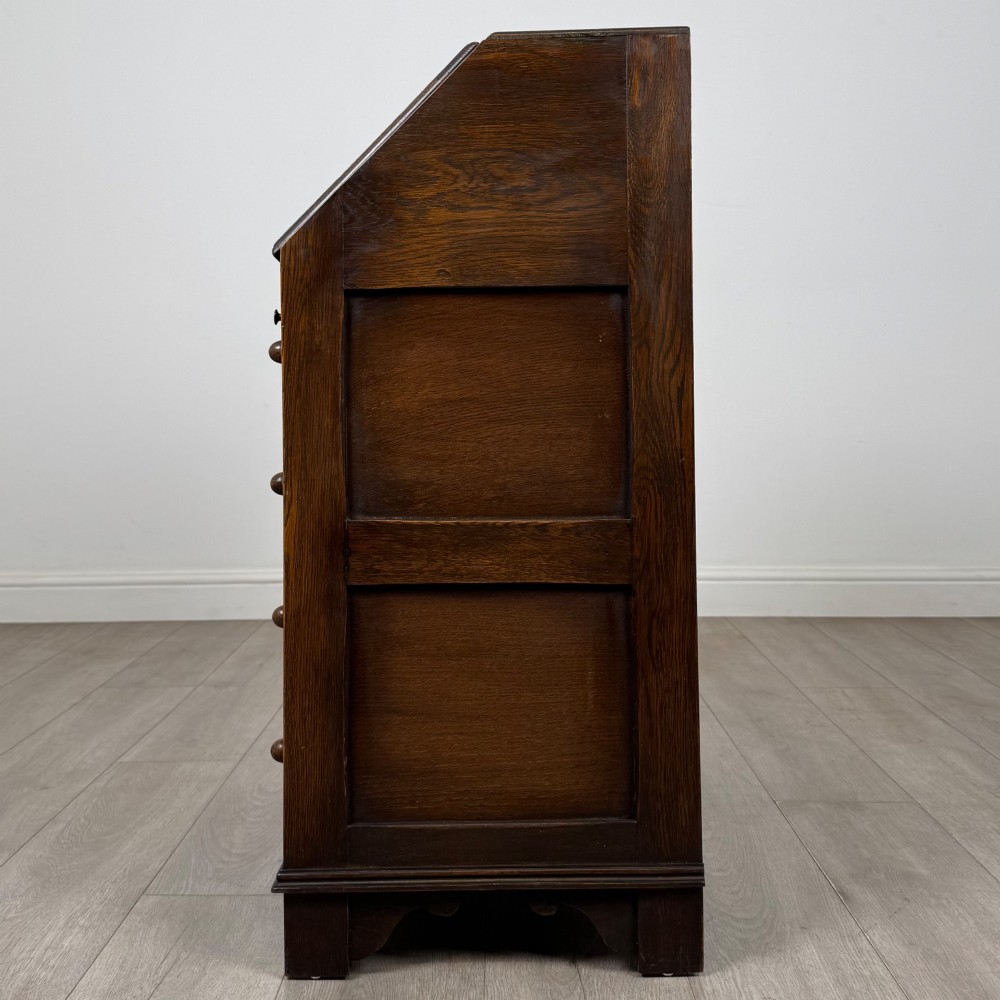 A dark oak writing bureau with a slanted fall-front upper section featuring three carved linenfold panels above four drawers with turned wooden knob handles. View IV