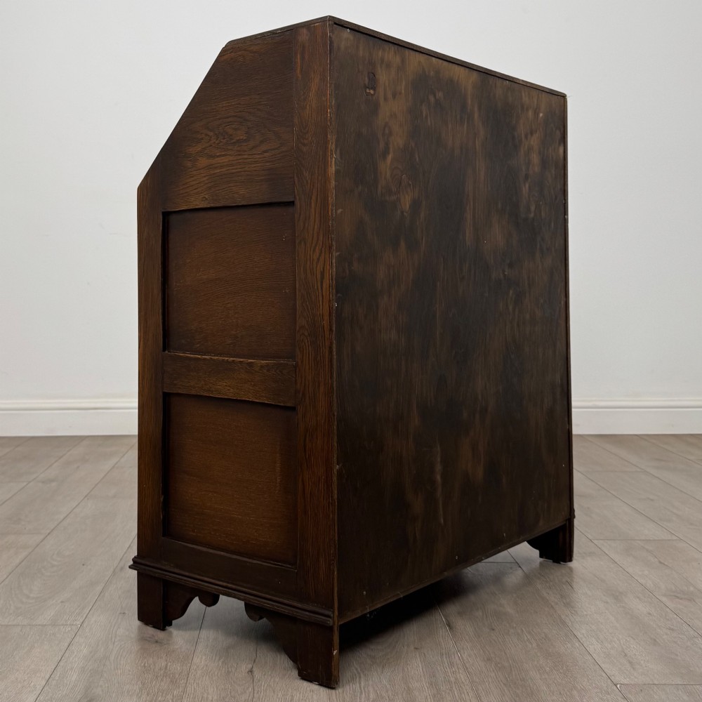 A dark oak writing bureau with a slanted fall-front upper section featuring three carved linenfold panels above four drawers with turned wooden knob handles. View V