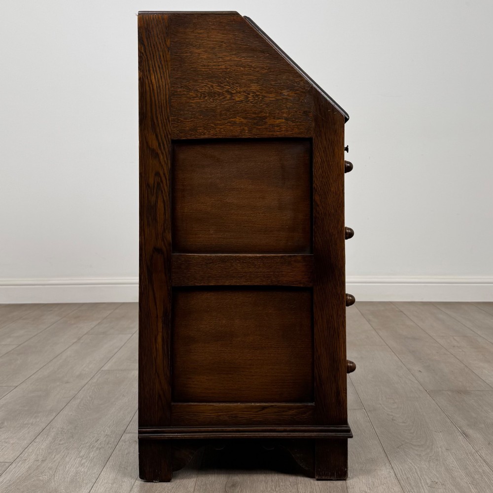 A dark oak writing bureau with a slanted fall-front upper section featuring three carved linenfold panels above four drawers with turned wooden knob handles. View VIII