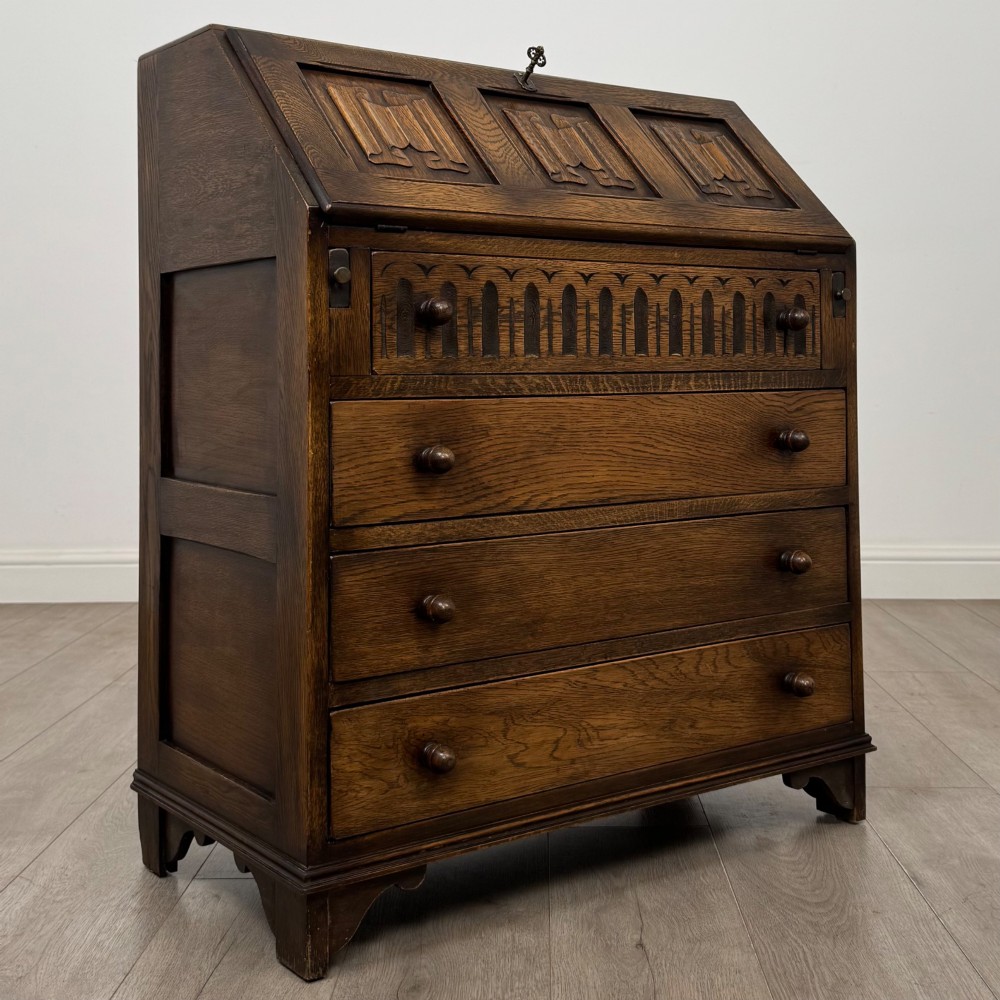 A dark oak writing bureau with a slanted fall-front upper section featuring three carved linenfold panels above four drawers with turned wooden knob handles. View IX