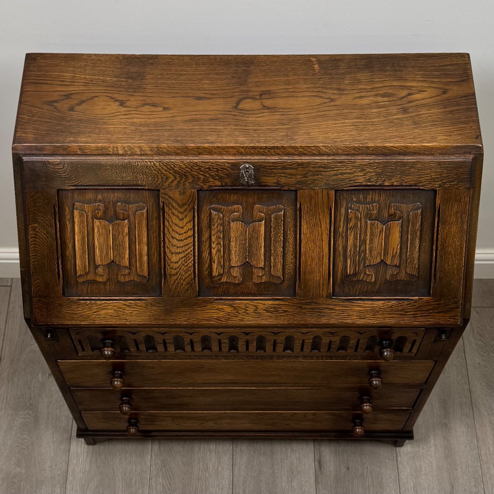 A dark oak writing bureau with a slanted fall-front upper section featuring three carved linenfold panels above four drawers with turned wooden knob handles. View X