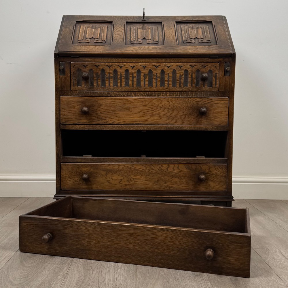 A dark oak writing bureau with a slanted fall-front upper section featuring three carved linenfold panels above four drawers with turned wooden knob handles. View XII