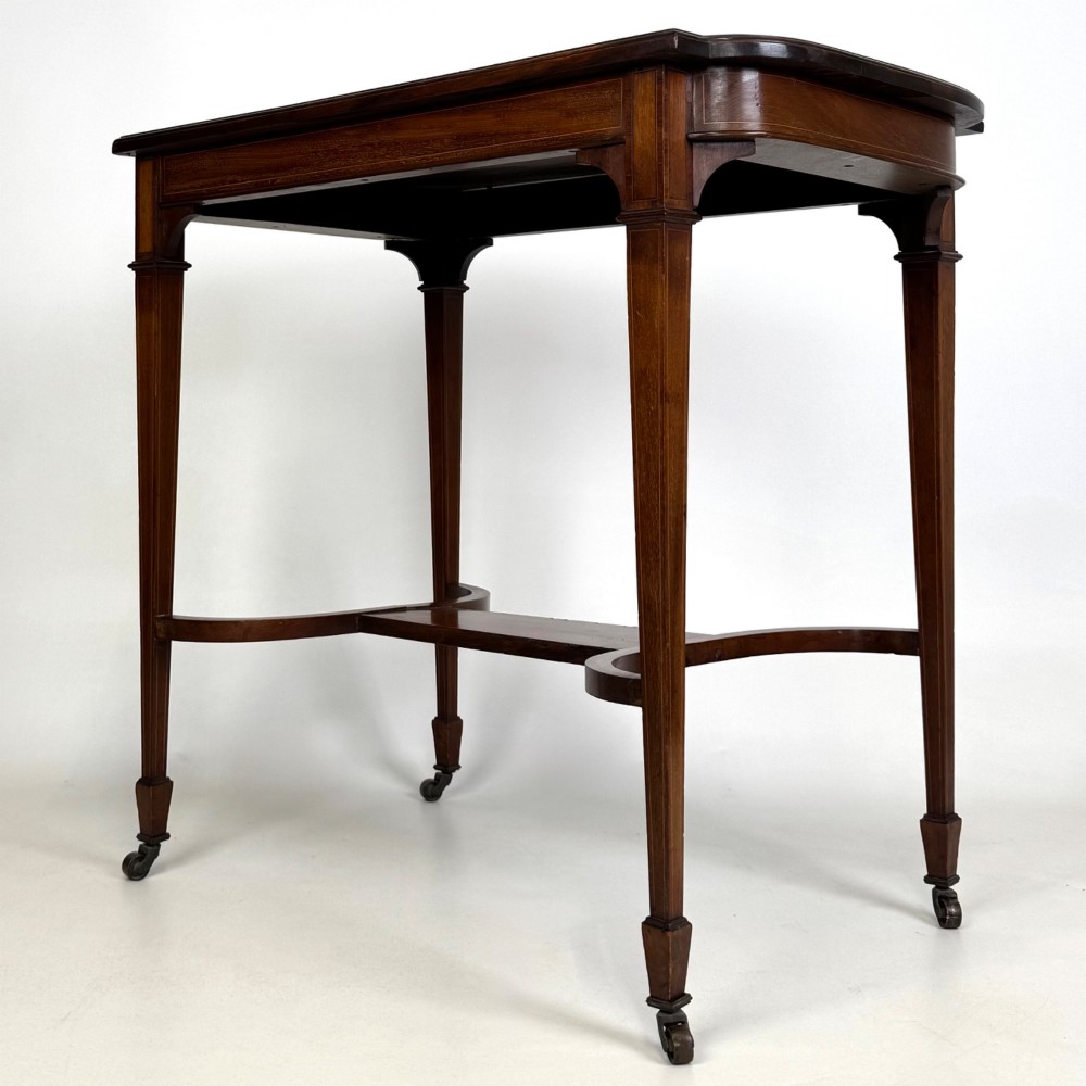 An early 20th-century mahogany side table featuring fine boxwood inlay along the apron, tapered square legs on brass castors, and a curved lower stretcher. View II