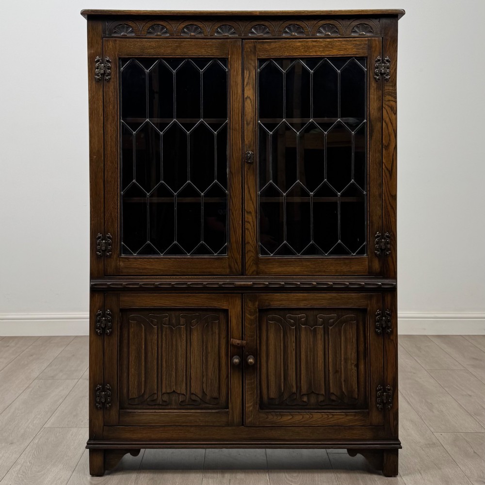 A dark oak display cabinet featuring two upper leaded glass doors, lower cupboard doors carved with linenfold panels, and decorative metal hinges.
