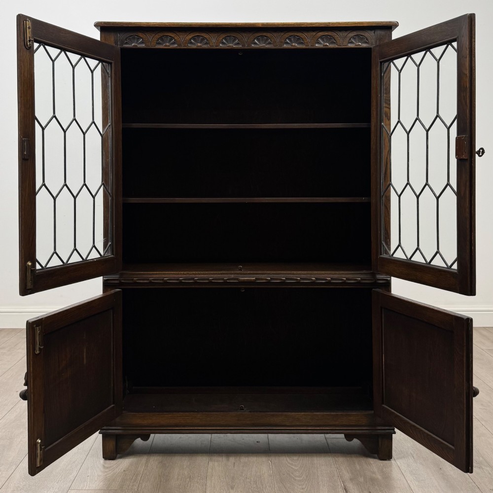 A dark oak display cabinet featuring two upper leaded glass doors, lower cupboard doors carved with linenfold panels, and decorative metal hinges. View II