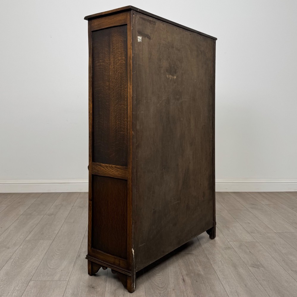 A dark oak display cabinet featuring two upper leaded glass doors, lower cupboard doors carved with linenfold panels, and decorative metal hinges. View V