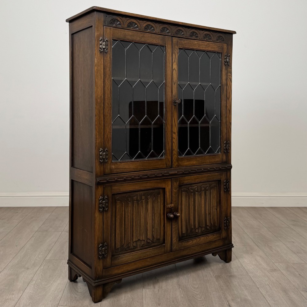 A dark oak display cabinet featuring two upper leaded glass doors, lower cupboard doors carved with linenfold panels, and decorative metal hinges. View IX