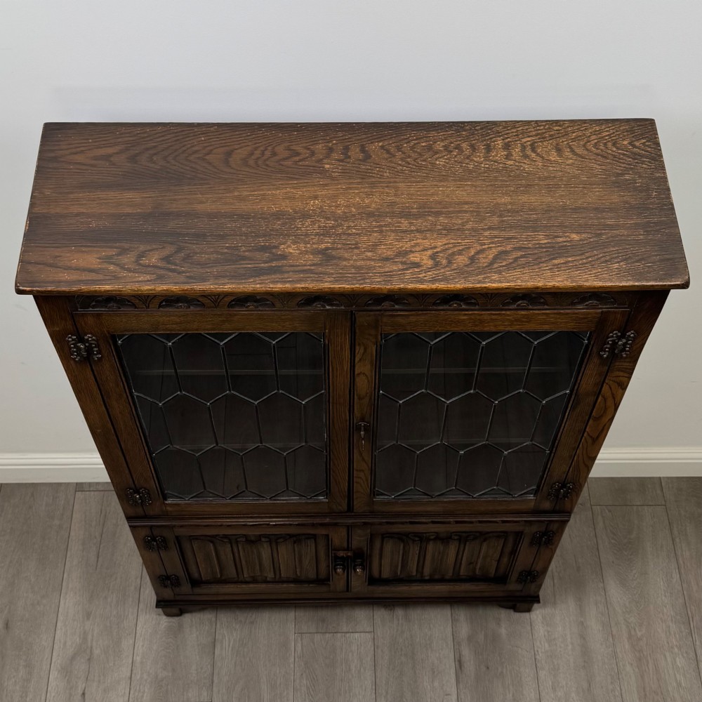 A dark oak display cabinet featuring two upper leaded glass doors, lower cupboard doors carved with linenfold panels, and decorative metal hinges. View X