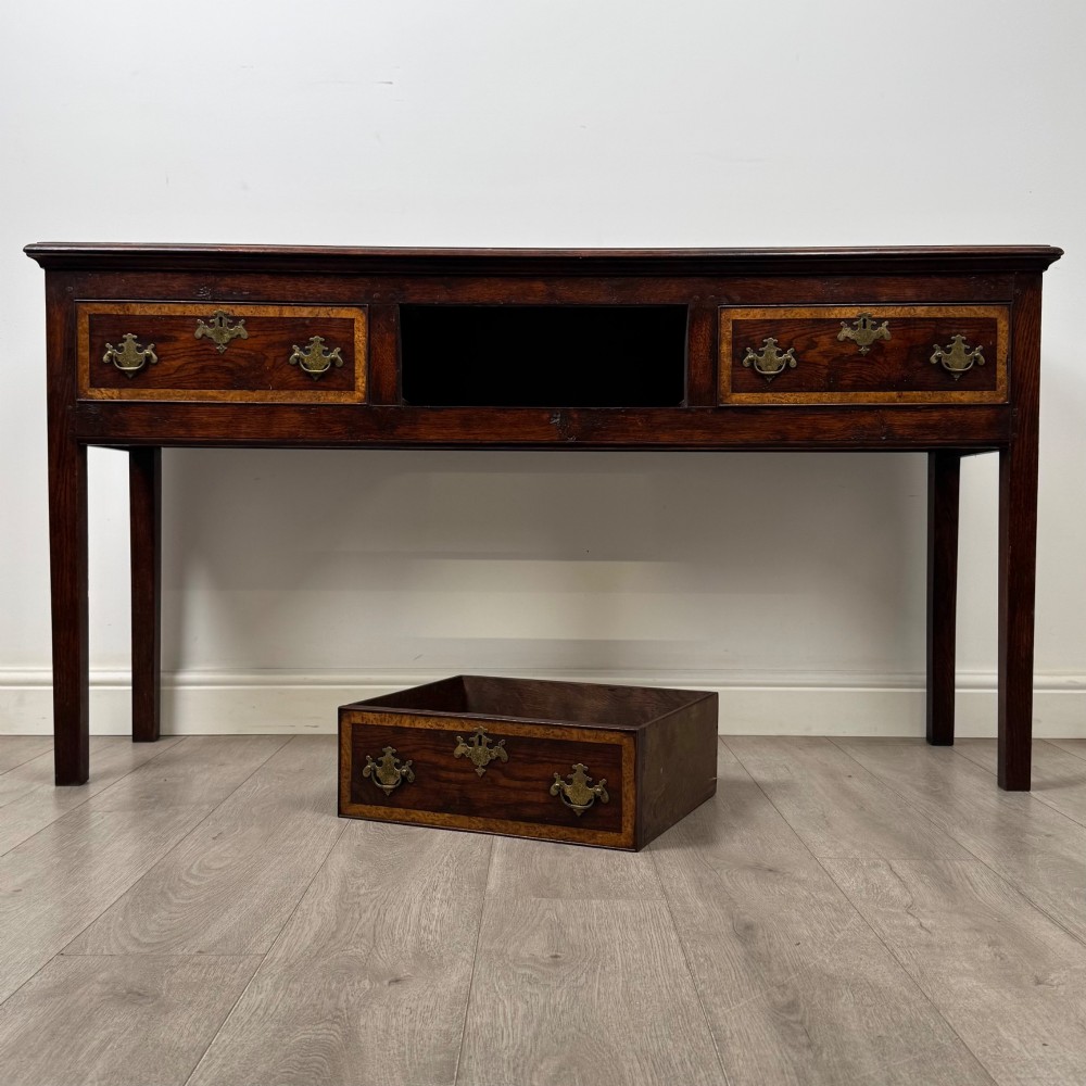 An antique dark oak dresser base featuring three crossbanded drawers with brass drop handles and keyhole escutcheons, supported by four straight square legs. View X