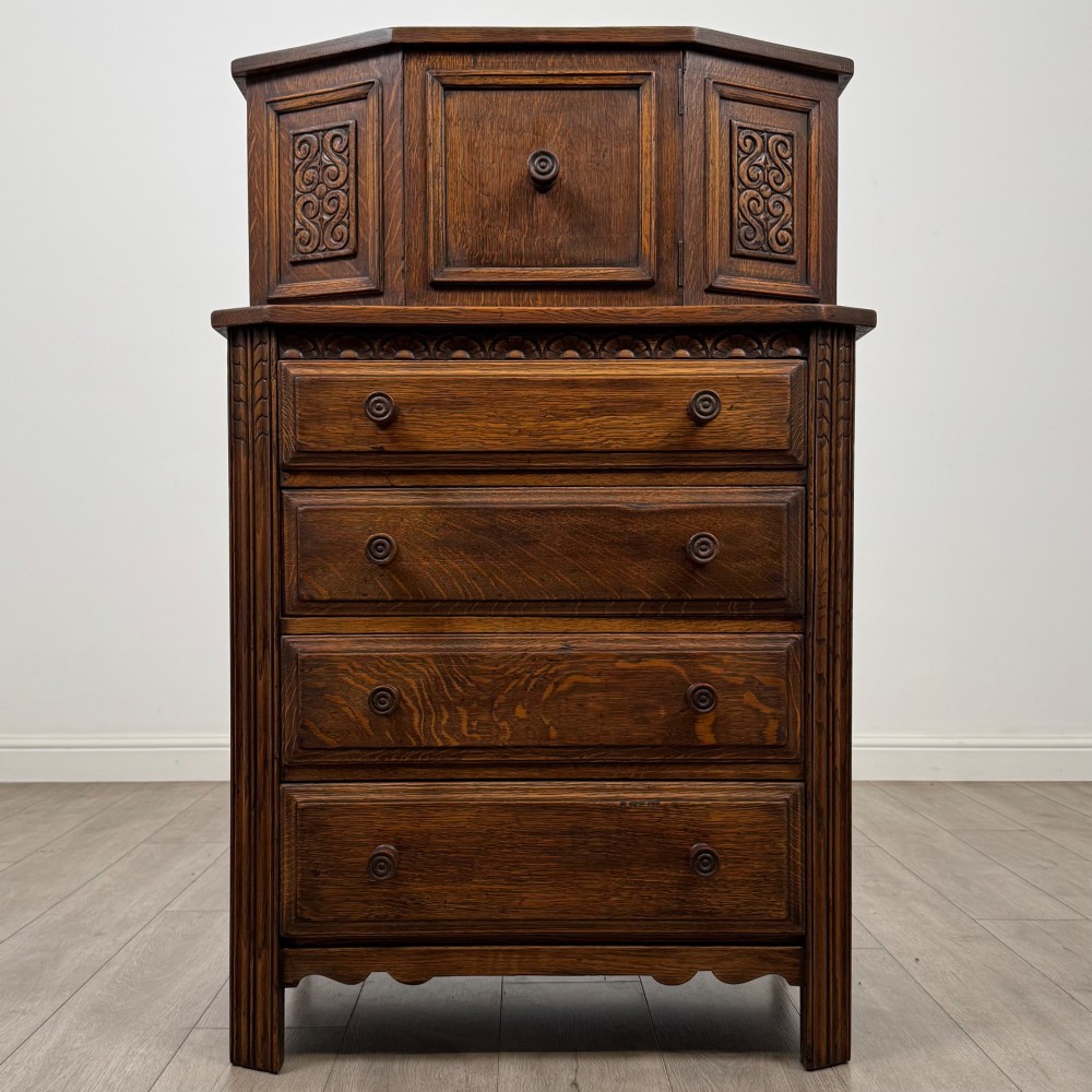 An Edwardian dark oak cupboard featuring an upper canted cabinet with carved decorative panels above four graduated drawers with turned wooden knobs.