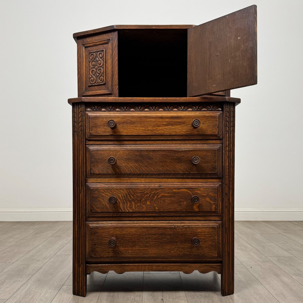 An Edwardian dark oak cupboard featuring an upper canted cabinet with carved decorative panels above four graduated drawers with turned wooden knobs. View II