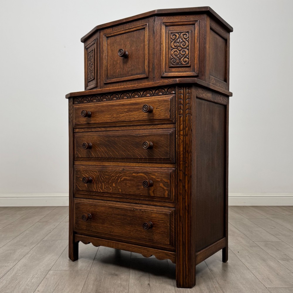 An Edwardian dark oak cupboard featuring an upper canted cabinet with carved decorative panels above four graduated drawers with turned wooden knobs. View III
