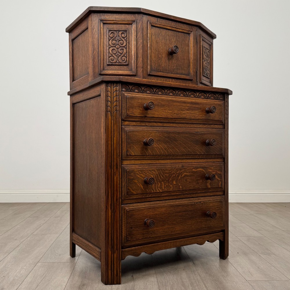 An Edwardian dark oak cupboard featuring an upper canted cabinet with carved decorative panels above four graduated drawers with turned wooden knobs. View IX