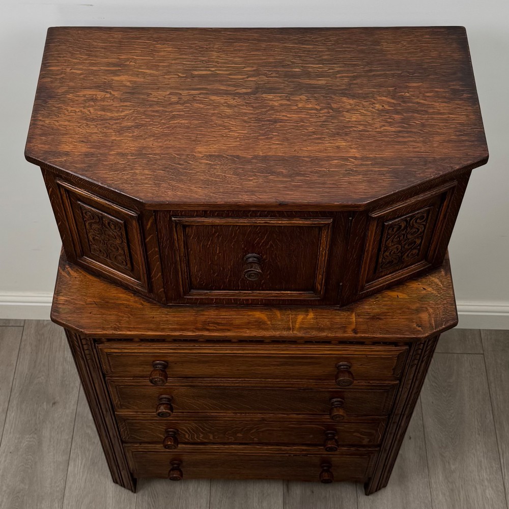 An Edwardian dark oak cupboard featuring an upper canted cabinet with carved decorative panels above four graduated drawers with turned wooden knobs. View X