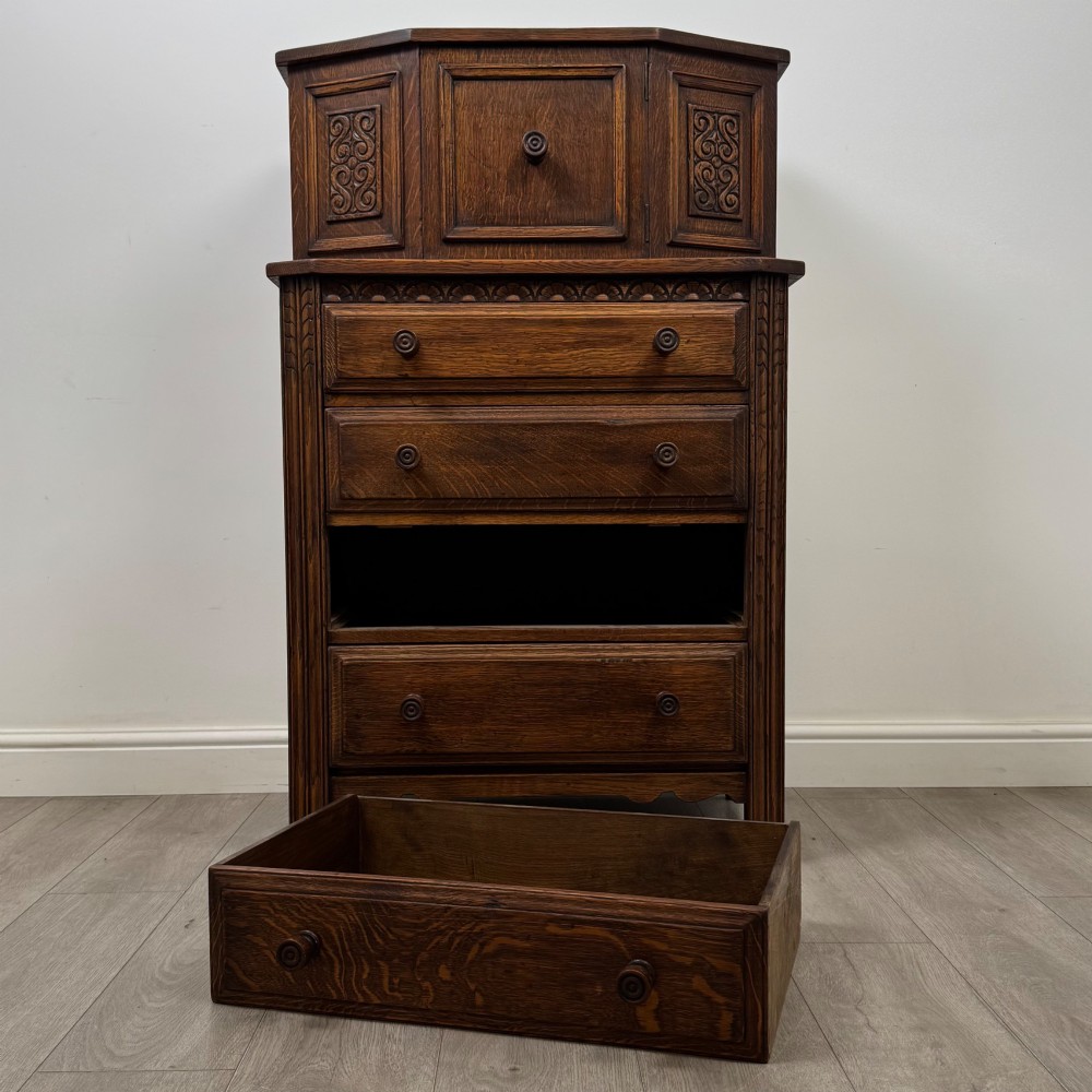 An Edwardian dark oak cupboard featuring an upper canted cabinet with carved decorative panels above four graduated drawers with turned wooden knobs. View XI