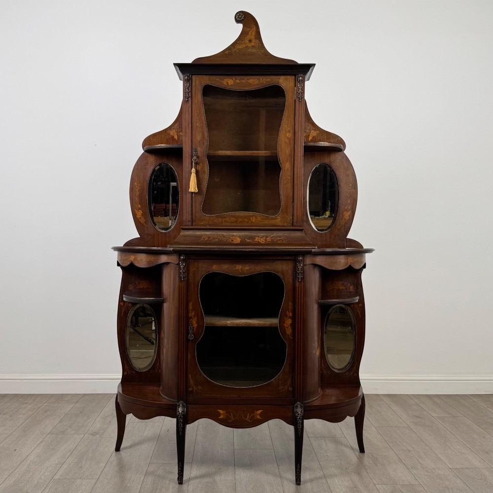 A late 19th-century mahogany display cabinet featuring floral marquetry inlay, glazed central doors, oval side mirrors, and a decorative asymmetric crest pediment.