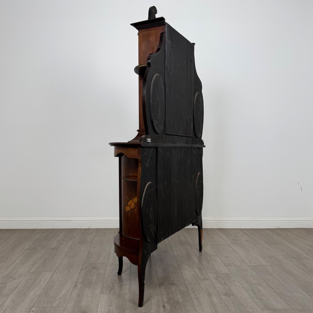 A late 19th-century mahogany display cabinet featuring floral marquetry inlay, glazed central doors, oval side mirrors, and a decorative asymmetric crest pediment. View V