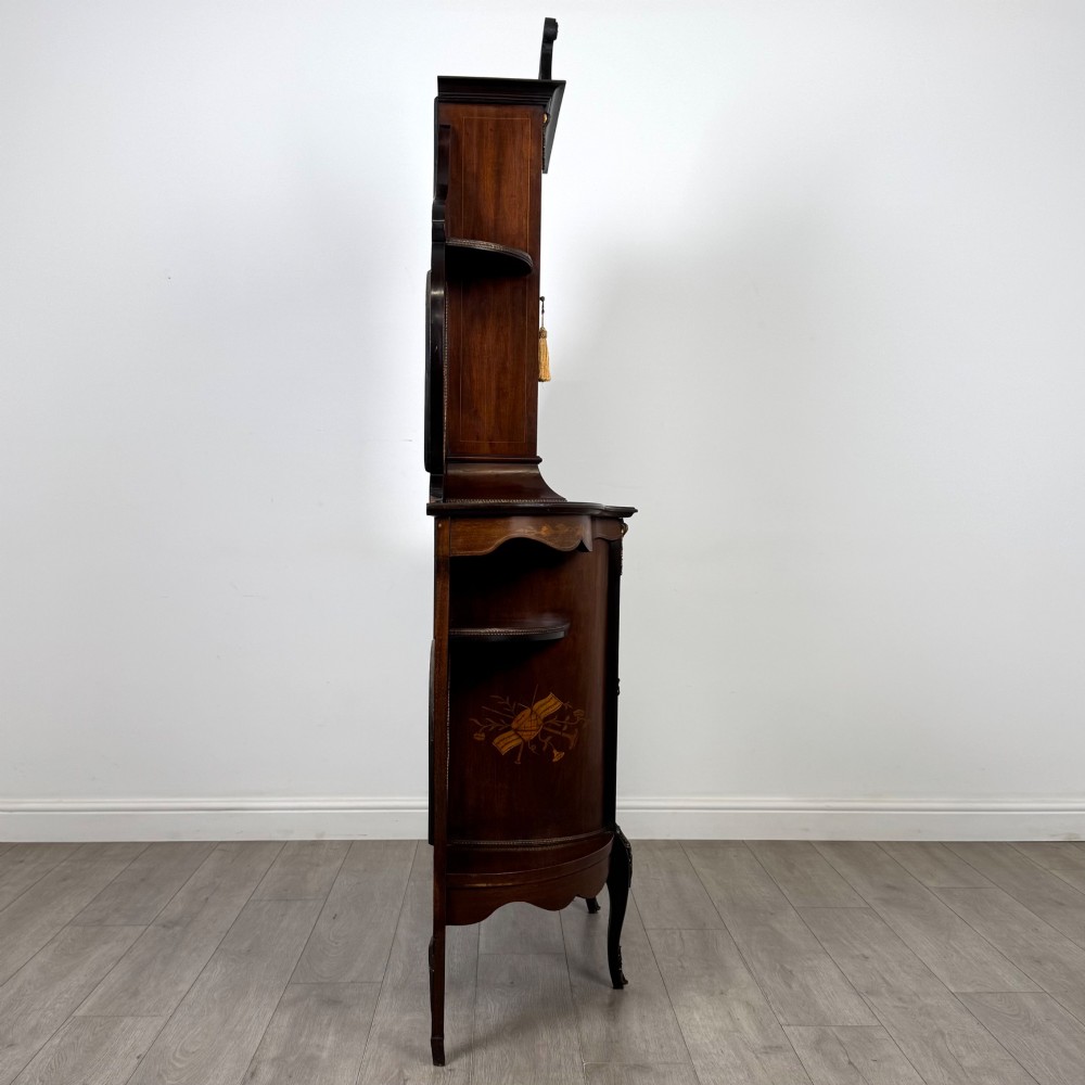A late 19th-century mahogany display cabinet featuring floral marquetry inlay, glazed central doors, oval side mirrors, and a decorative asymmetric crest pediment. View VIII