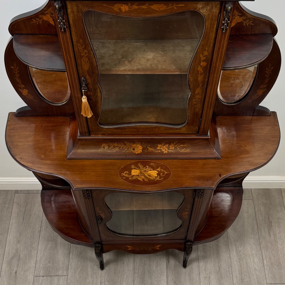 A late 19th-century mahogany display cabinet featuring floral marquetry inlay, glazed central doors, oval side mirrors, and a decorative asymmetric crest pediment. View X
