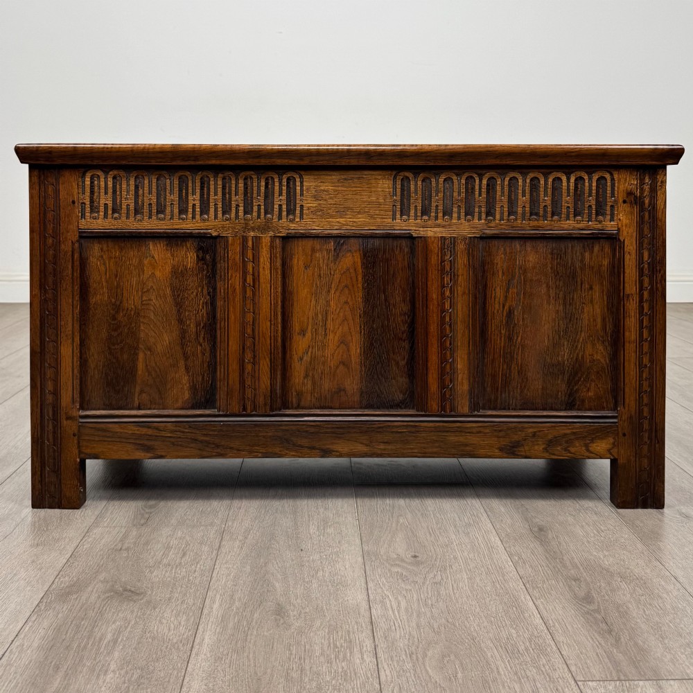 A rectangular dark oak blanket box featuring a hinged top lid, panelled front sections, and carved arch motifs along the upper frieze.