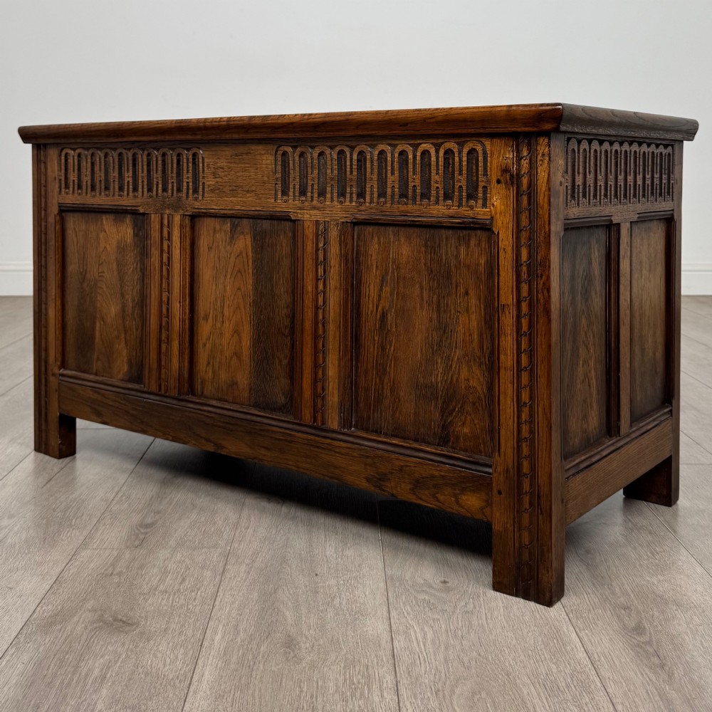 A rectangular dark oak blanket box featuring a hinged top lid, panelled front sections, and carved arch motifs along the upper frieze. View II