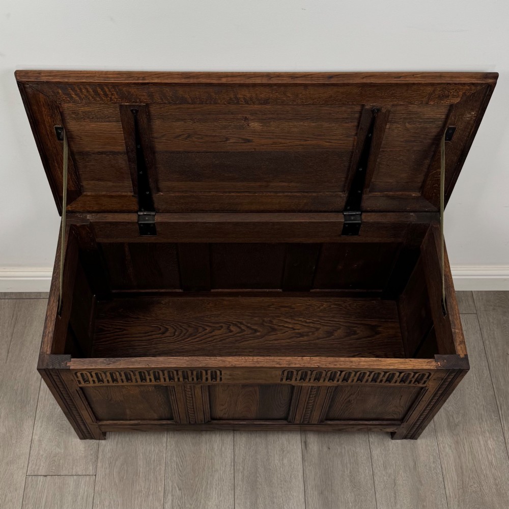 A rectangular dark oak blanket box featuring a hinged top lid, panelled front sections, and carved arch motifs along the upper frieze. View X