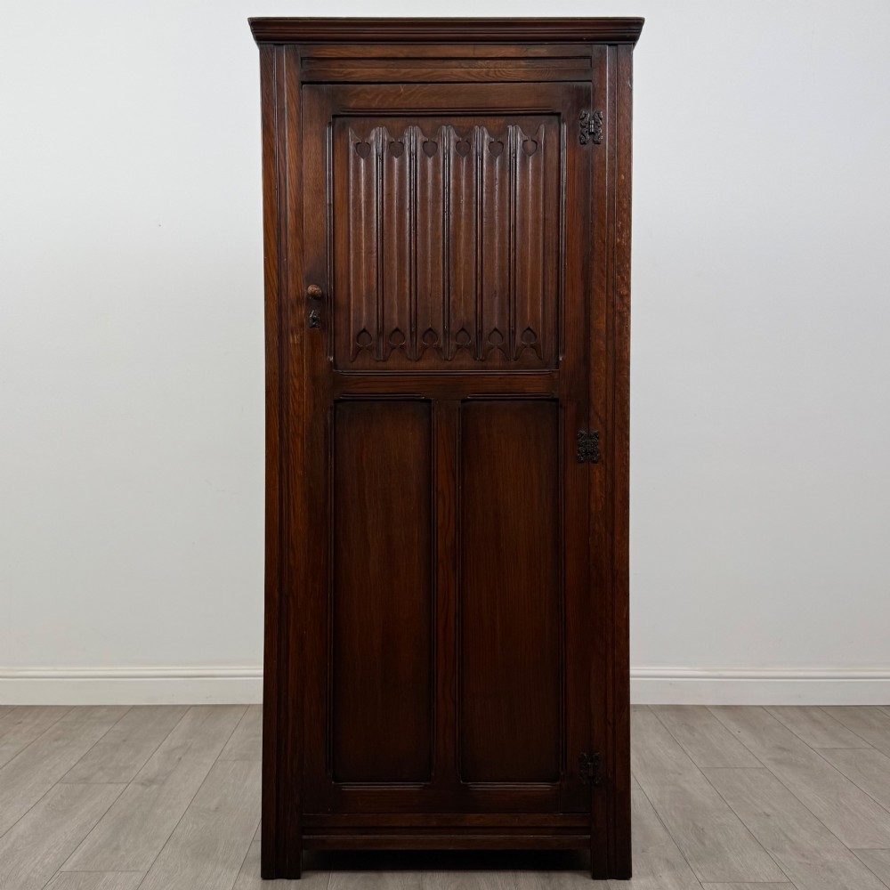 A tall single-door dark oak Edwardian wardrobe featuring a carved upper linenfold panel and decorative iron hinges.