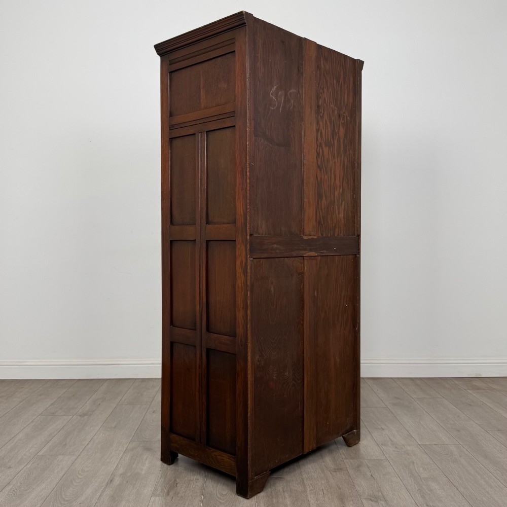 A tall single-door dark oak Edwardian wardrobe featuring a carved upper linenfold panel and decorative iron hinges. View V