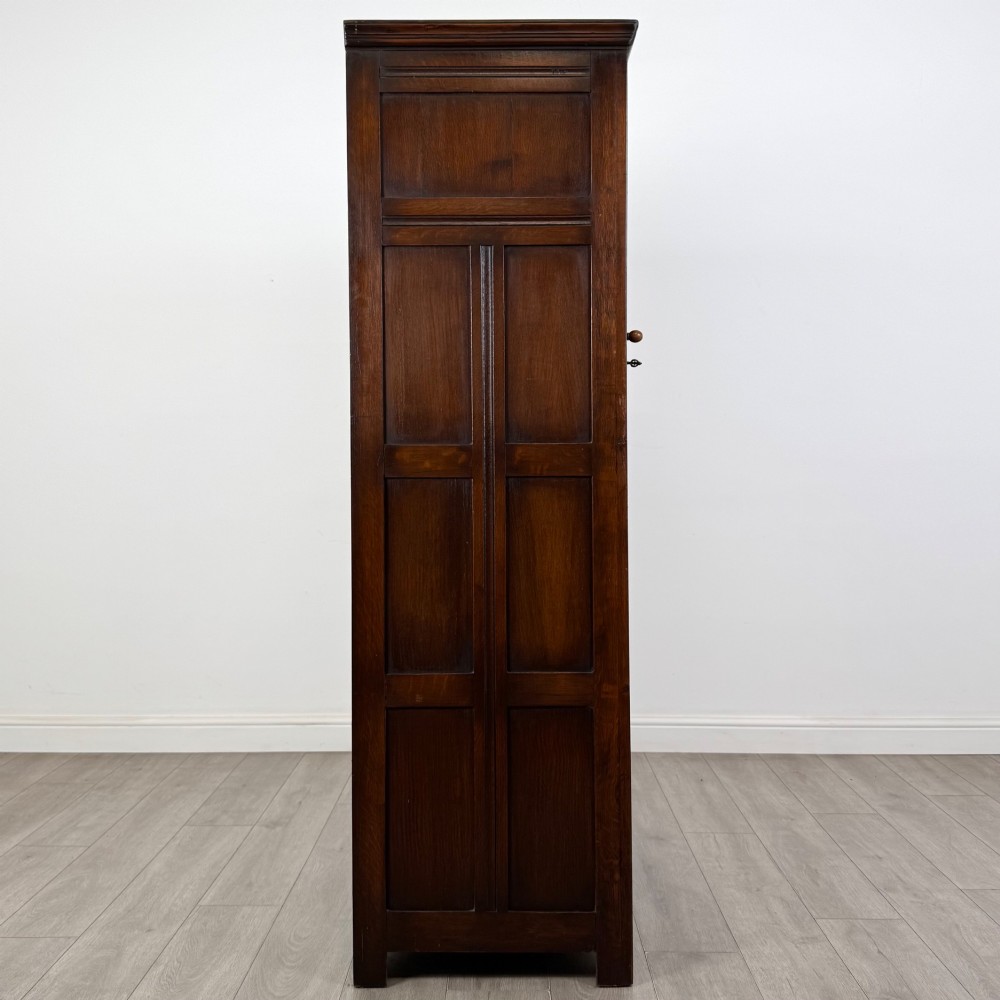 A tall single-door dark oak Edwardian wardrobe featuring a carved upper linenfold panel and decorative iron hinges. View VIII