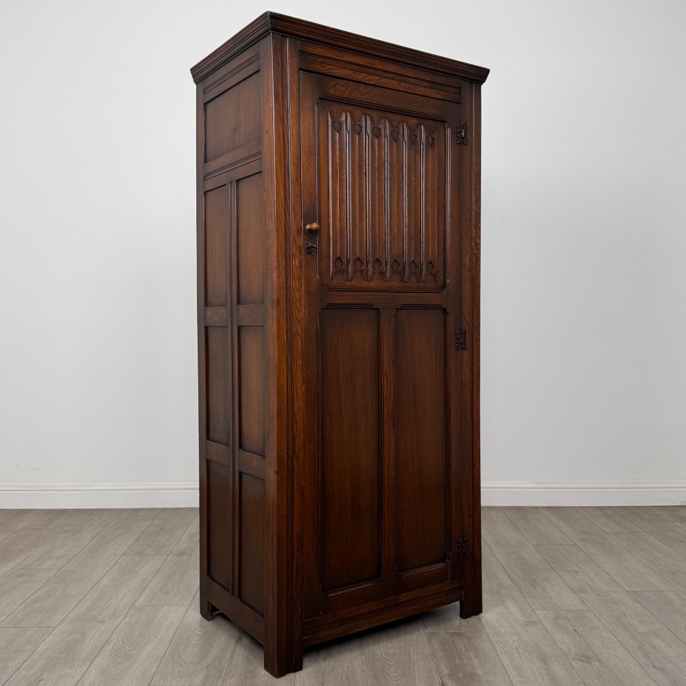 A tall single-door dark oak Edwardian wardrobe featuring a carved upper linenfold panel and decorative iron hinges. View IX