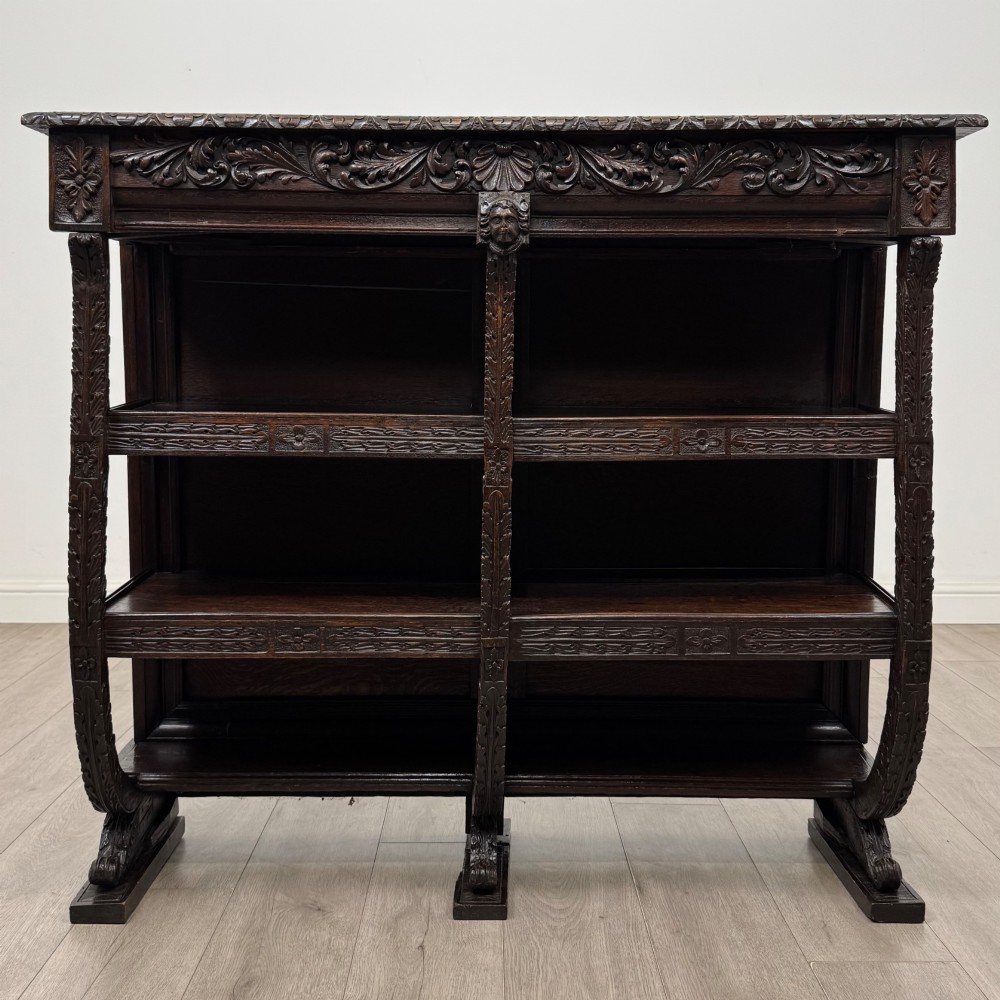 A 19th-century dark oak open bookcase featuring hand-carved frieze foliage, a central mask motif, and three tiers of open shelving.