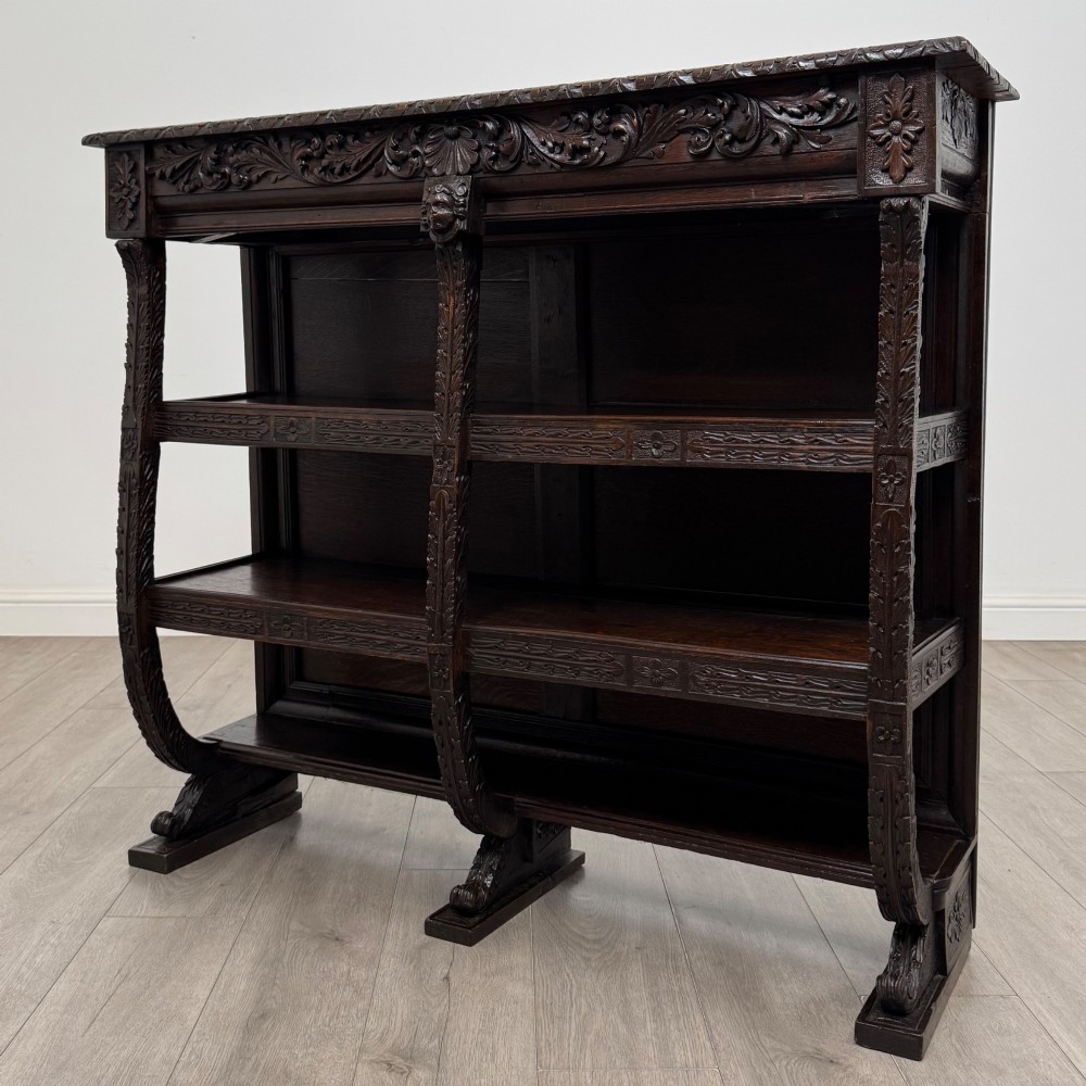 A 19th-century dark oak open bookcase featuring hand-carved frieze foliage, a central mask motif, and three tiers of open shelving. View II