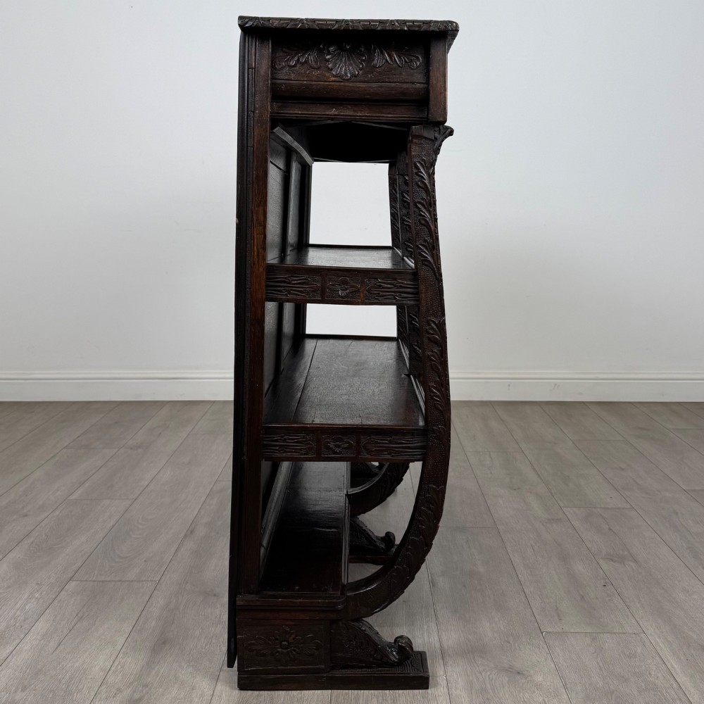 A 19th-century dark oak open bookcase featuring hand-carved frieze foliage, a central mask motif, and three tiers of open shelving. View VII
