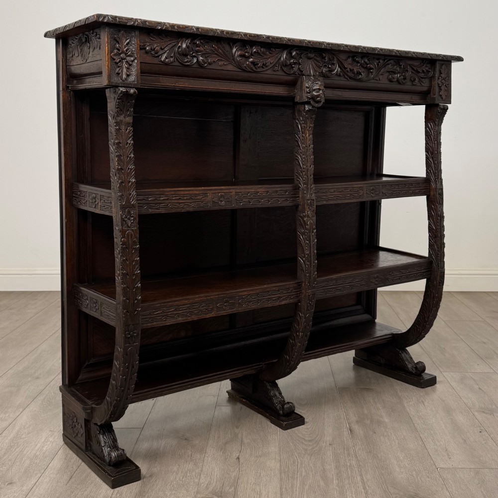 A 19th-century dark oak open bookcase featuring hand-carved frieze foliage, a central mask motif, and three tiers of open shelving. View VIII