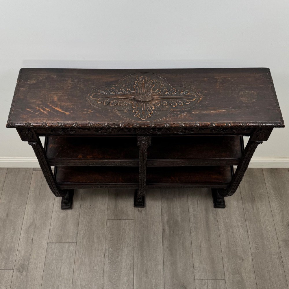 A 19th-century dark oak open bookcase featuring hand-carved frieze foliage, a central mask motif, and three tiers of open shelving. View IX